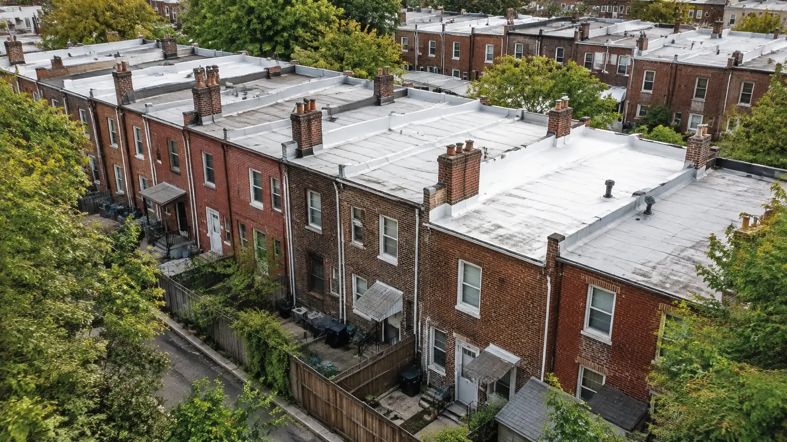 Rear low-slope roofs of a row of brick rowhomes covered in white and gray membrane, with chimneys and parapet walls